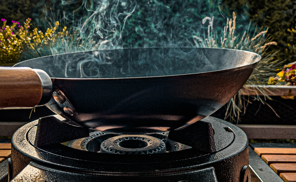 Smoke rising from a carbon steel wok with a wooden handle outdoors